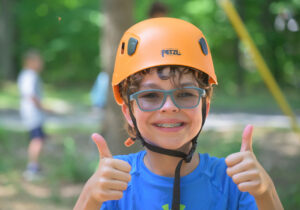 A child wearing an orange helmet and glasses gives two thumbs up while smiling outdoors.